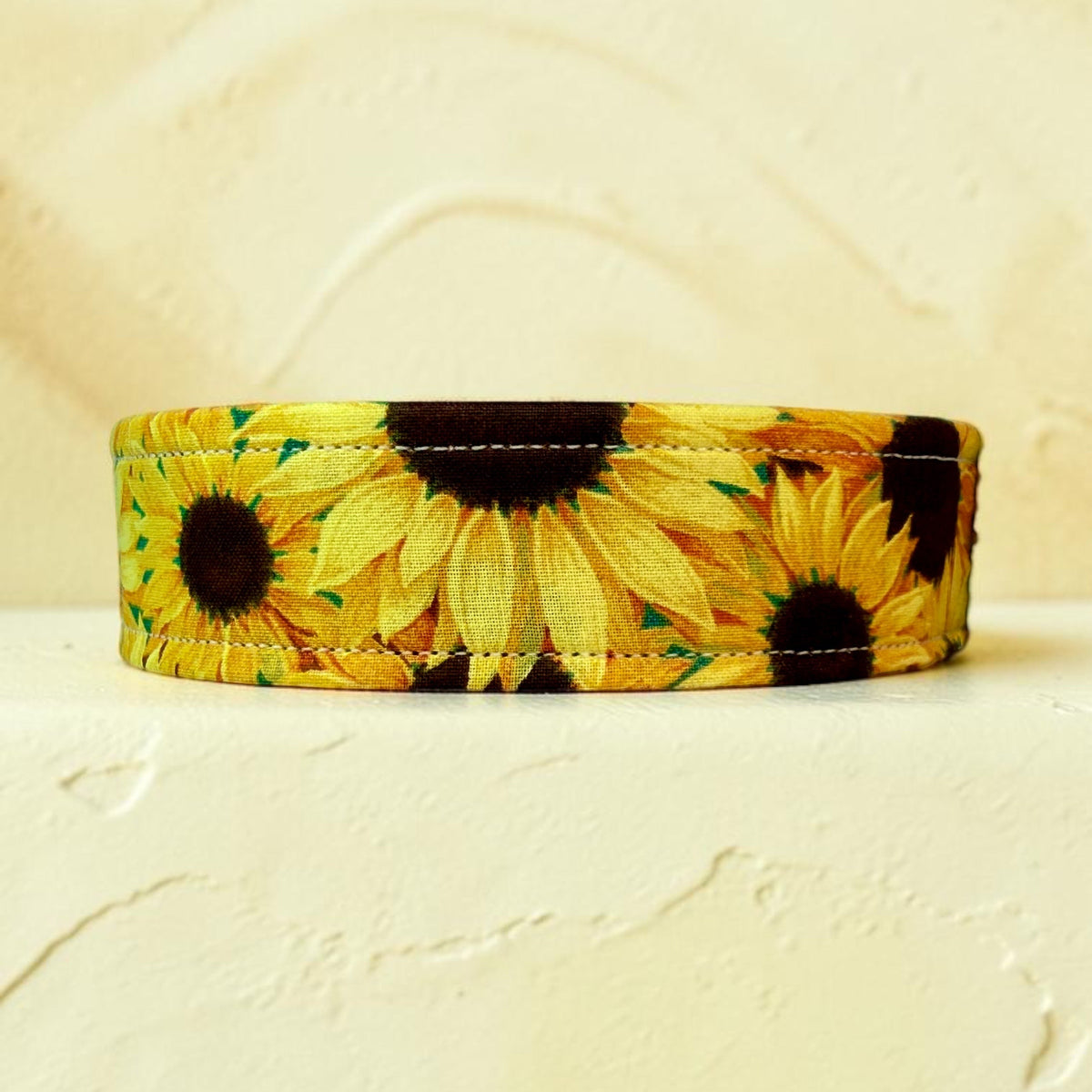 A fabric dog collar featuring a vibrant yellow and brown sunflower print, displayed on a textured cream-colored surface.